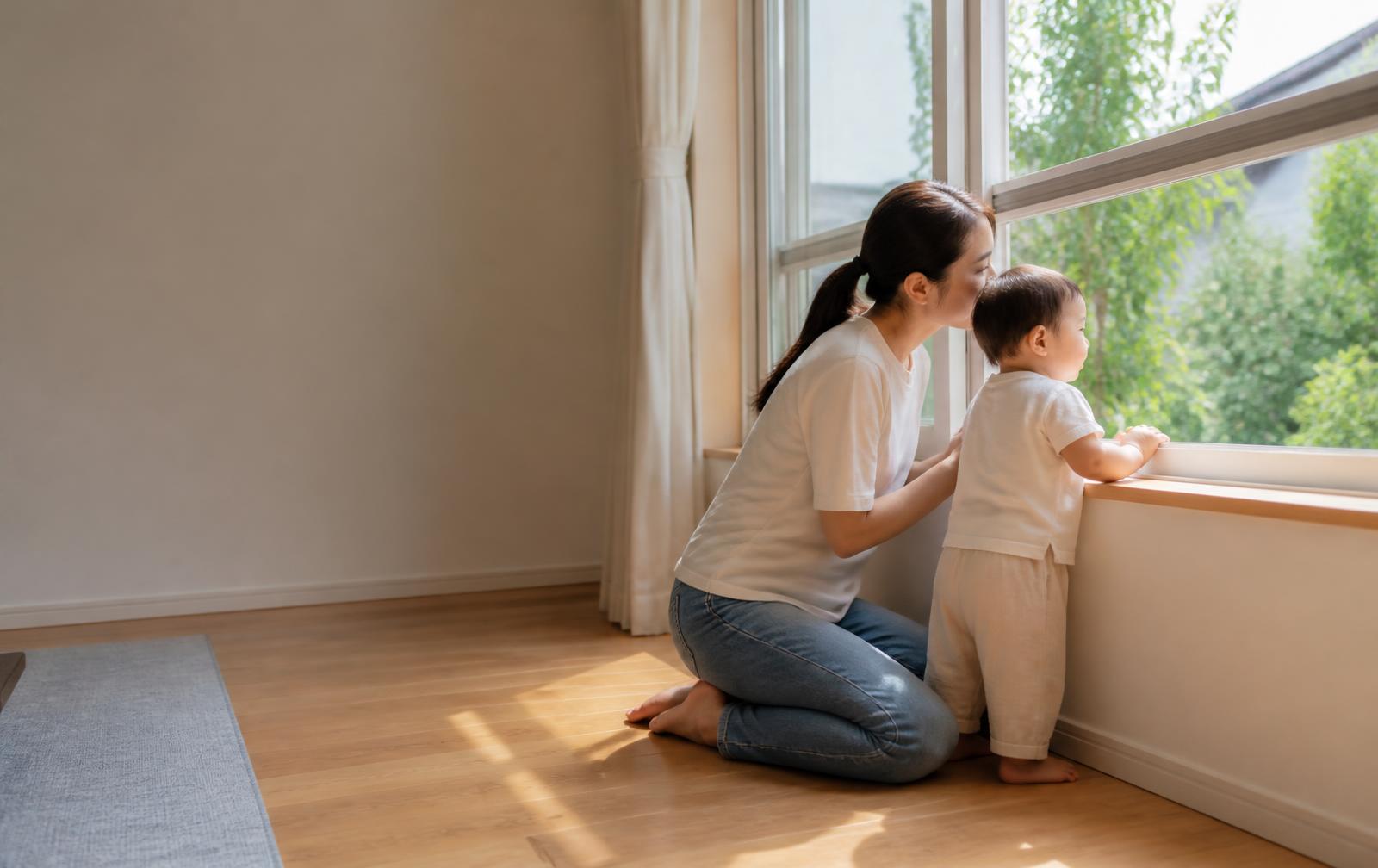 A mother kneeling beside her toddler as they look out of a sunlit window in an empty room